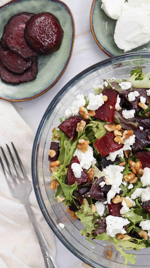 A glass bowl of fall salad with beets, featuring mixed greens, roasted beets, goat cheese, and walnuts. Nearby, a plate holds beet slices and another has a mound of goat cheese. A fork and napkin are placed beside the bowl.