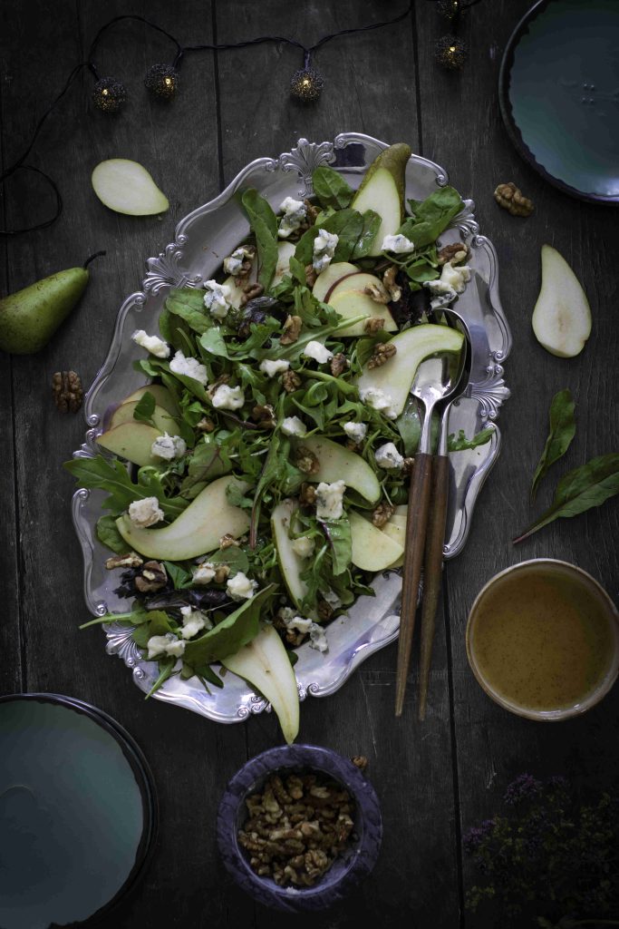 An overhead view of an Easy Pear and Walnut Salad with crumbled cheese on a silver tray, surrounded by salad ingredients and dishes on a dark wooden table—an inviting Fall Harvest Salad Recipe that celebrates seasonal flavors.