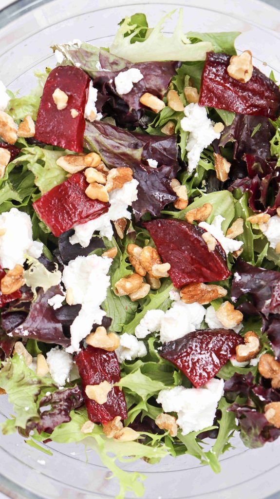 A close-up of a fall salad with beets, featuring mixed greens, roasted beet slices, crumbled goat cheese, and walnut pieces in a glass bowl.