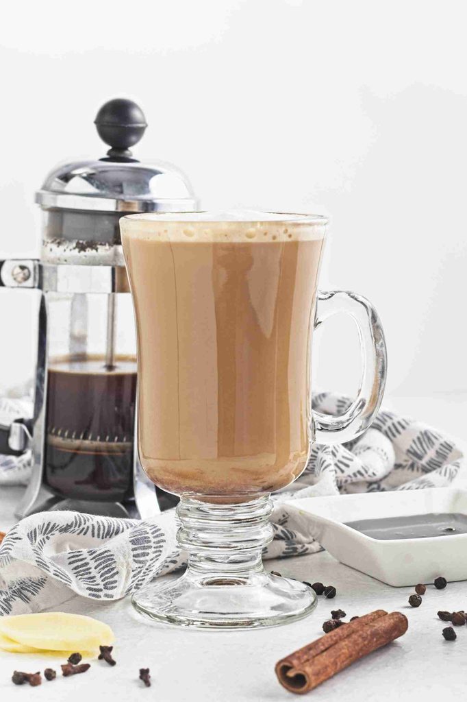 A glass mug of chai latte sits on a white surface next to a French press, a cinnamon stick, whole spices, and a white dish, with a patterned cloth in the background. The cozy scene is elevated by the addition of gingerbread syrup, infusing the drink with warm, spiced sweetness.