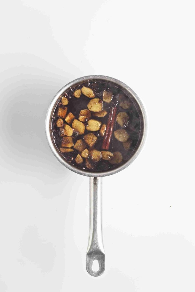 A saucepan filled with sliced ginger, cinnamon sticks, and star anise simmering in dark liquid—perfect for making gingerbread syrup—is viewed from above on a white background.
