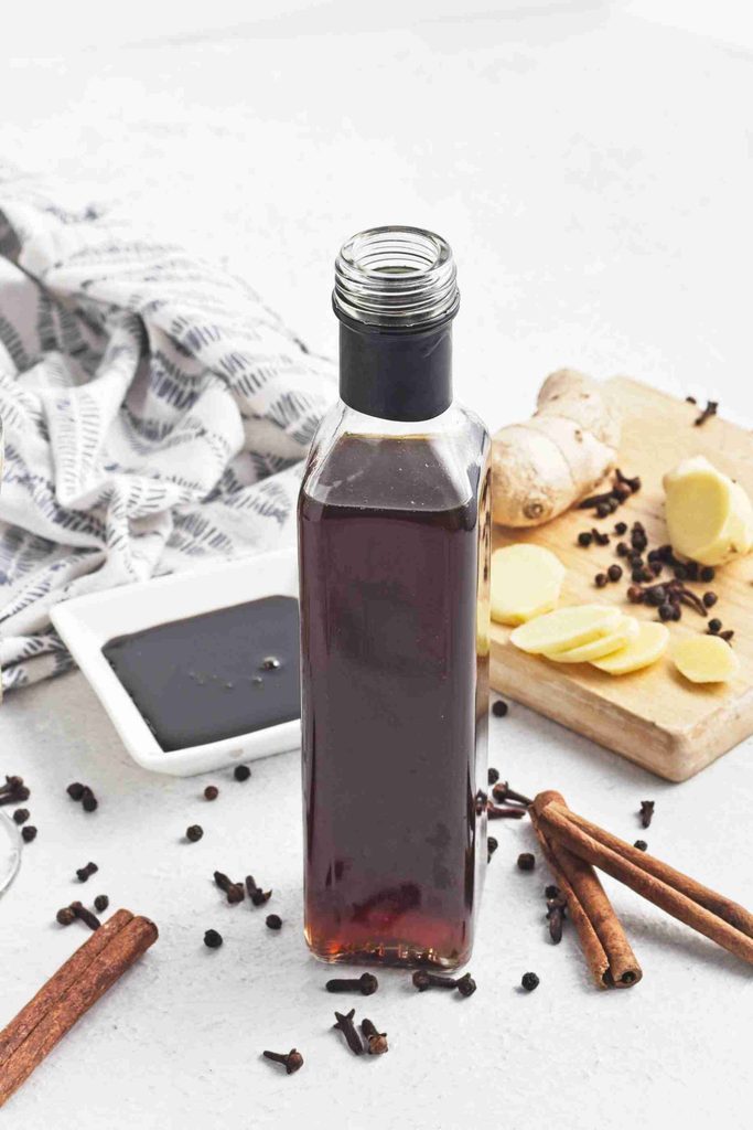 A bottle of dark liquid, likely gingerbread syrup, is surrounded by cinnamon sticks, cloves, fresh ginger slices on a cutting board, and a small dish of sauce, all set on a white surface with a patterned cloth in the background.