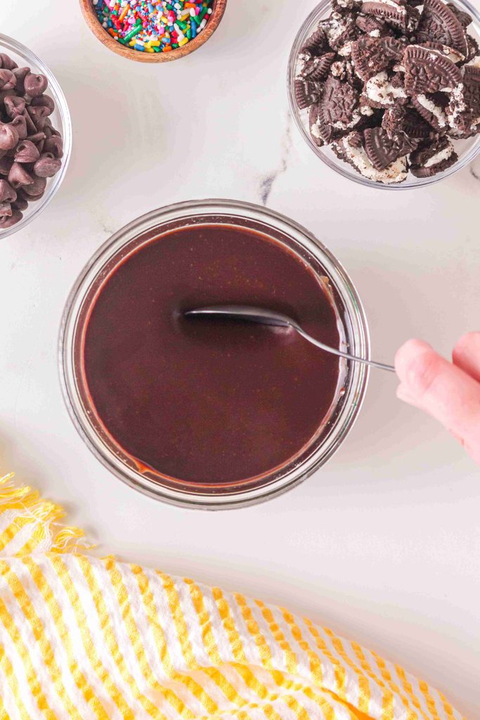 A hand stirs chocolate sauce in a bowl, surrounded by bowls of sprinkles, crushed cookies, and chocolate chips, with a yellow striped cloth nearby—perfect for preparing a homemade hot fudge sauce or trying your favorite hot fudge sauce recipe.