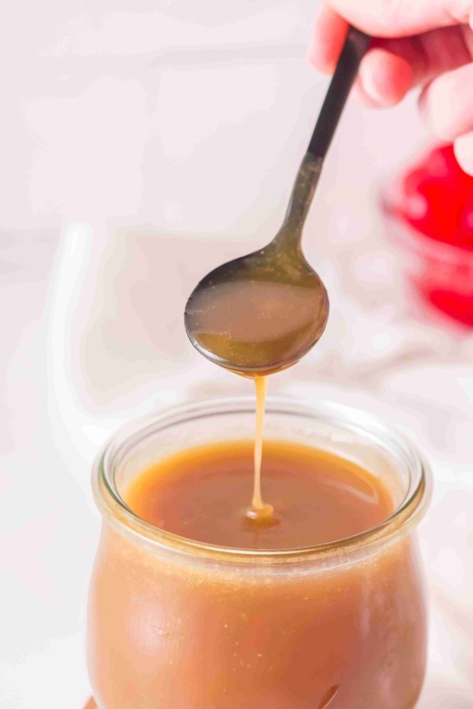 A spoon drizzling caramel sauce back into a glass jar filled with caramel, showcasing the perfect texture for any butterscotch sauce recipe, with a blurred background and a hand holding the spoon.