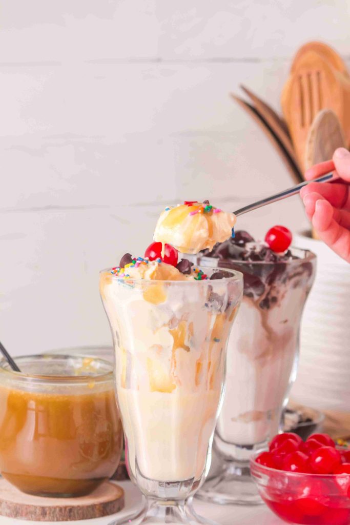 A hand holds a spoonful of ice cream above a glass sundae topped with whipped cream, sprinkles, and a cherry. Another sundae, a jar of rich butterscotch sauce—perfect for trying your favorite butterscotch sauce recipe—and a bowl of cherries are in the background.