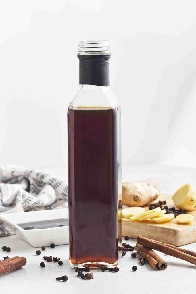 A bottle of dark gingerbread syrup sits on a white surface, surrounded by cinnamon sticks, peppercorns, sliced ginger, and a patterned cloth, with a ginger root and spices on a wooden board in the background.