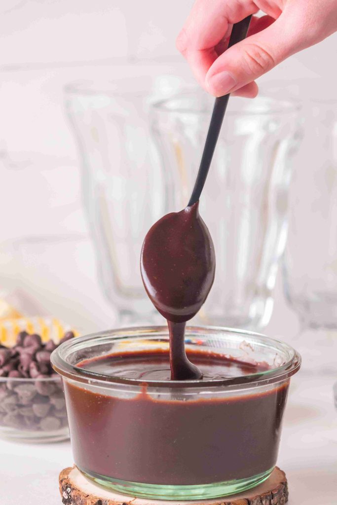 A hand holds a spoon dripping chocolate sauce over a glass bowl filled with rich homemade hot fudge sauce. Blurred glassware and chocolate chips are in the background, hinting at an indulgent hot fudge sauce recipe made with real chocolate chips.