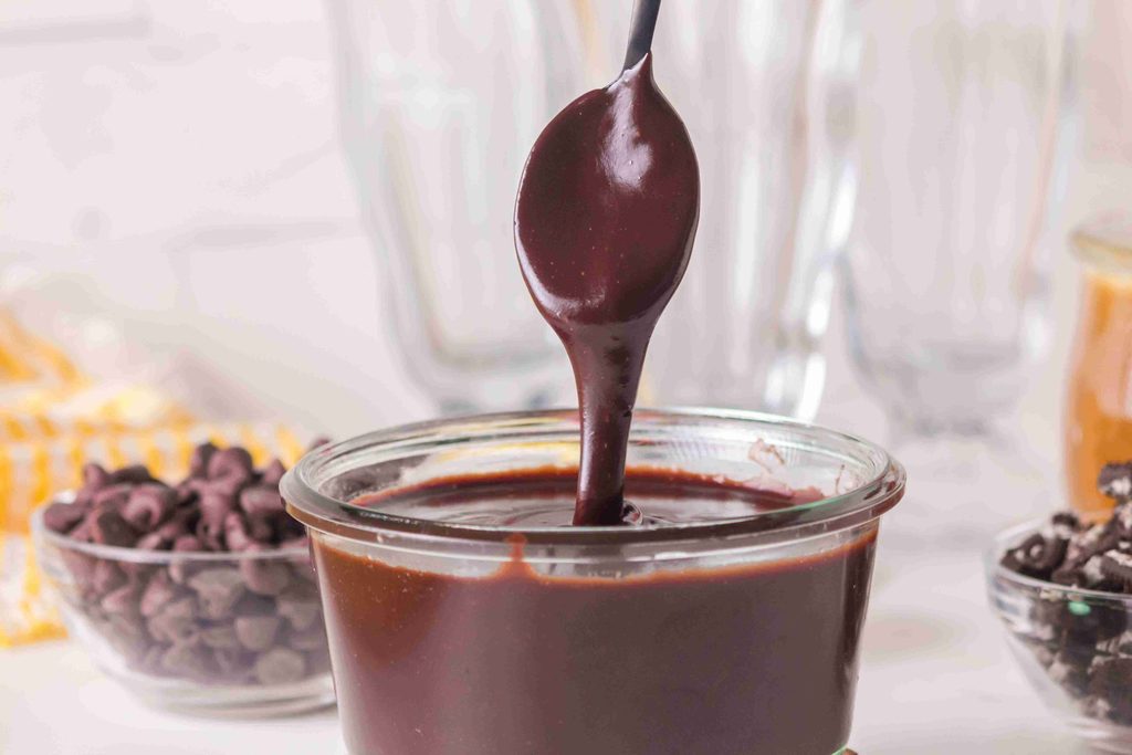 A spoon drips thick homemade hot fudge sauce back into a small glass jar, with chocolate chips and dessert ingredients blurred in the background.