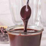 A spoon drips thick homemade hot fudge sauce back into a small glass jar, with chocolate chips and dessert ingredients blurred in the background.