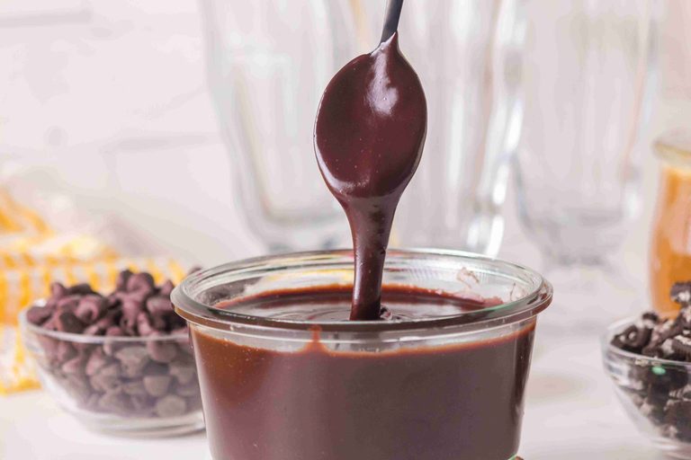 A spoon drips thick homemade hot fudge sauce back into a small glass jar, with chocolate chips and dessert ingredients blurred in the background.