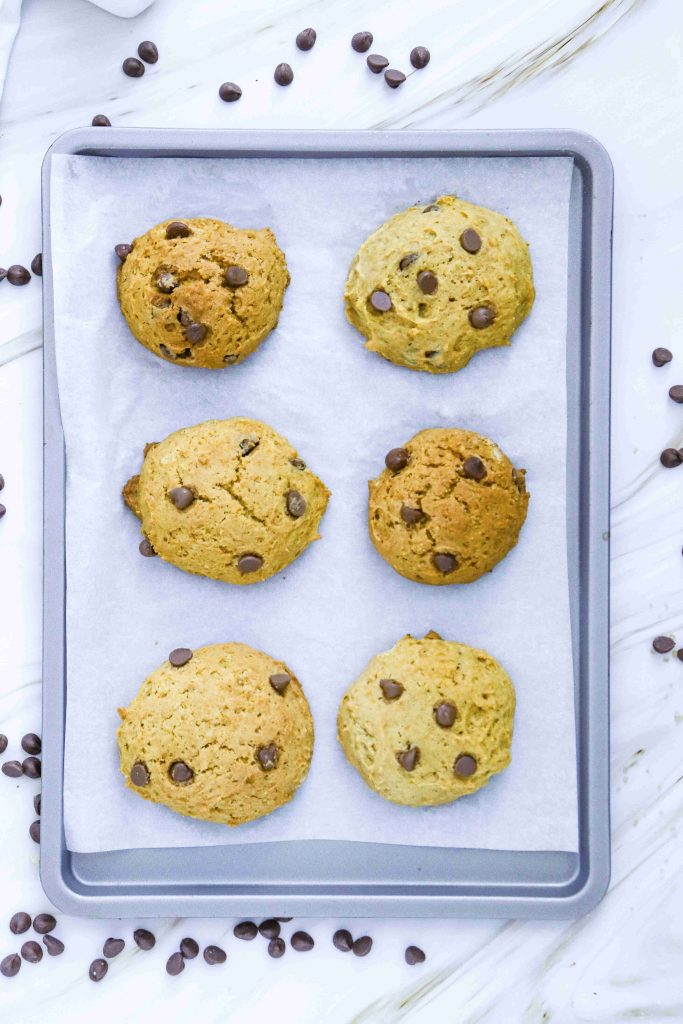 Six chocolate chip cookies on a parchment-lined baking sheet, with chocolate chips scattered around on a white marble surface—perfect inspiration for anyone craving pumpkin cookies with chocolate chips or searching for a soft pumpkin cookie recipe.