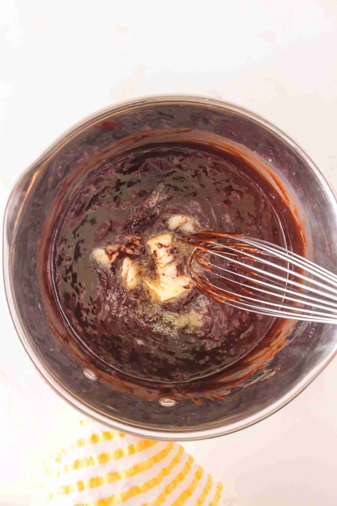 A saucepan filled with chocolate mixture, being whisked with melted butter in the center—just the start of a delicious homemade hot fudge sauce. A metal whisk is inside the pot, and a yellow-striped towel is partially visible at the bottom.