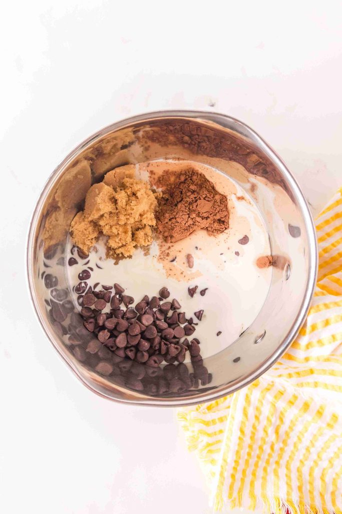 A metal bowl with chocolate chips, brown sugar, cocoa powder, and cream—perfect for making a homemade hot fudge sauce—sits next to a yellow and white striped cloth on a white surface.