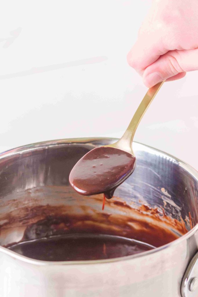A hand holds a spoon with thick homemade hot fudge sauce above a saucepan filled with more chocolatey goodness, showcasing an irresistible hot fudge sauce recipe perfect for drizzling over desserts.