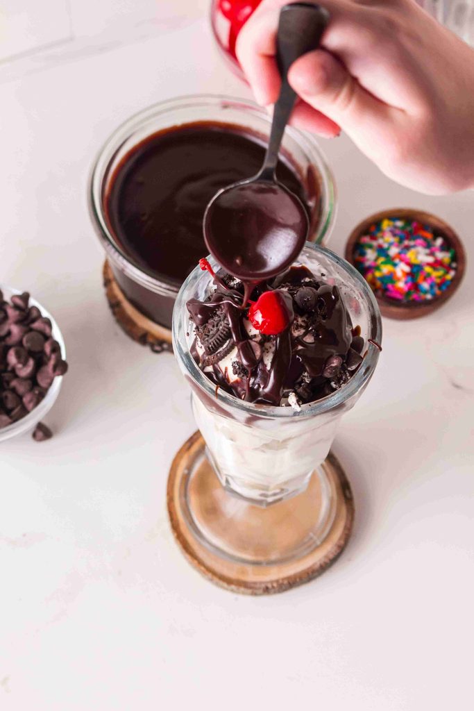 A hand drizzles homemade hot fudge sauce over a sundae topped with a cherry, chocolate pieces, and whipped cream, with sprinkles and chocolate chips nearby.