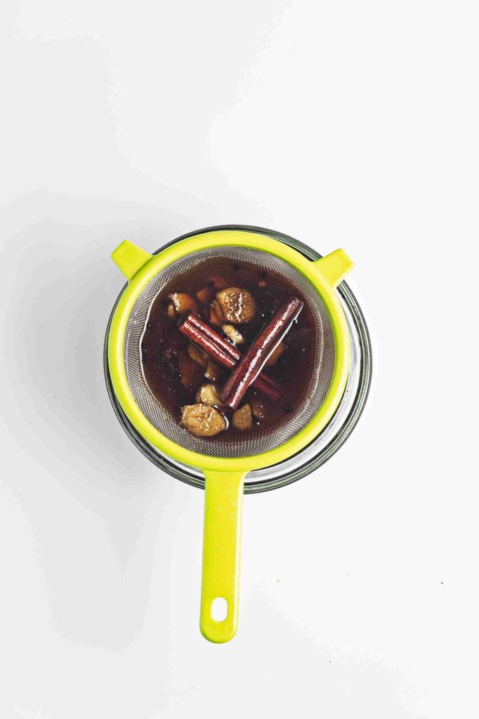 A yellow strainer with cinnamon sticks and ginger pieces sits over a jar, filtering a dark gingerbread syrup on a white background.