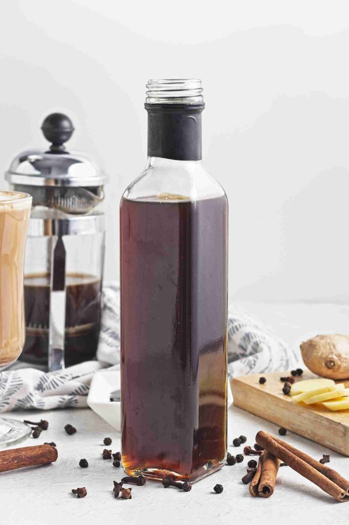 A glass bottle filled with dark liquid sits on a table near a French press, coffee, cinnamon sticks, ginger slices, black peppercorns, and a light cloth—perfect ingredients for preparing homemade gingerbread syrup.
