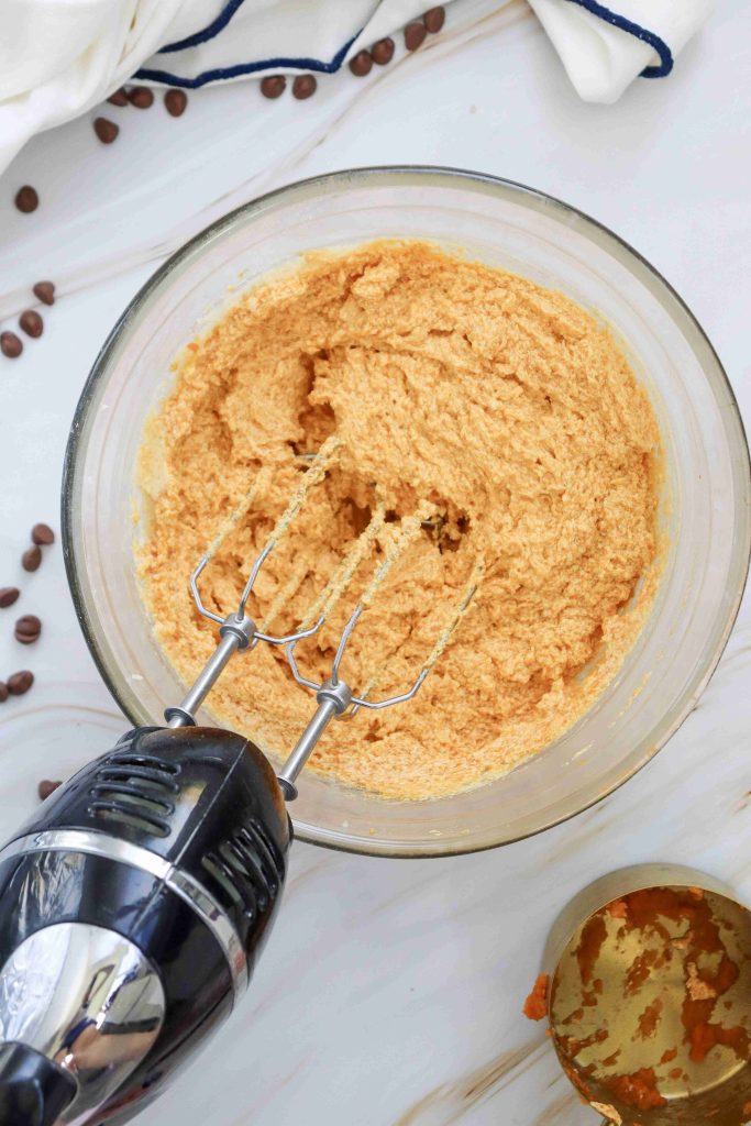 A glass bowl filled with a creamy, tan batter for soft pumpkin cookies with chocolate chips is being mixed with an electric hand mixer on a marble surface, with coffee beans scattered nearby.