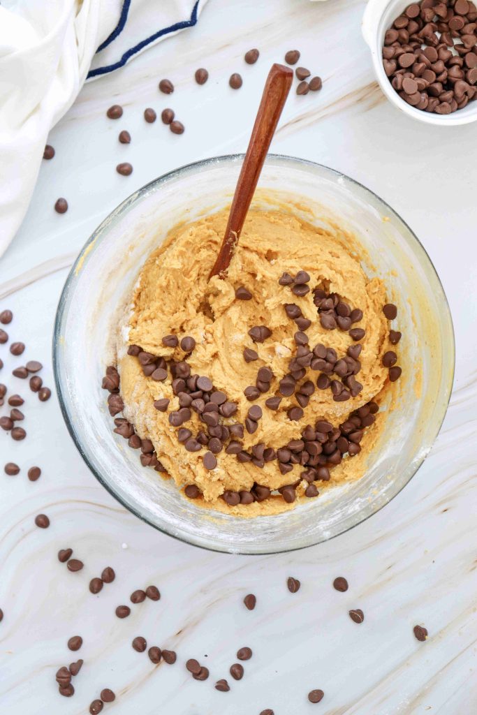 A glass bowl of cookie dough with chocolate chips and a wooden spoon, perfect for making soft pumpkin cookies with chocolate chips. Chocolate chips are scattered on the marble countertop around the bowl, capturing the cozy feel of a classic soft pumpkin cookie recipe.