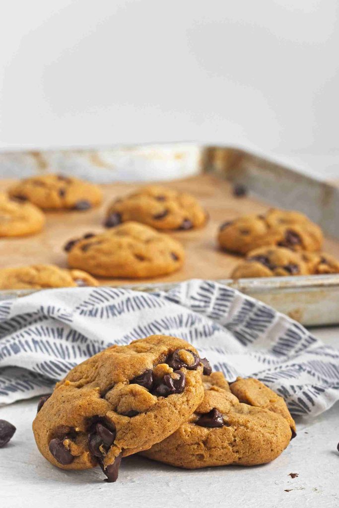 Two golden brown vegan pumpkin chocolate chip cookies rest on a patterned cloth in front of a baking tray with more cookies in the background. The cookies are studded with visible chocolate chips, showcasing their delicious plant-based twist.