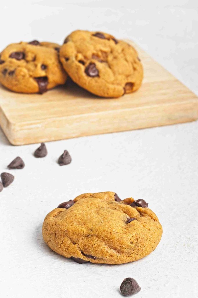 A vegan pumpkin chocolate chip cookie sits on a white surface with a few chocolate chips nearby. In the background, three more cookies rest on a wooden board.