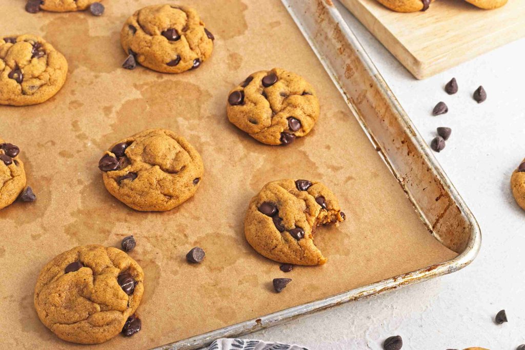 A baking tray with vegan pumpkin chocolate chip cookies on parchment paper. One cookie has a bite taken out. Some chocolate chips and cookies are scattered on the table nearby.