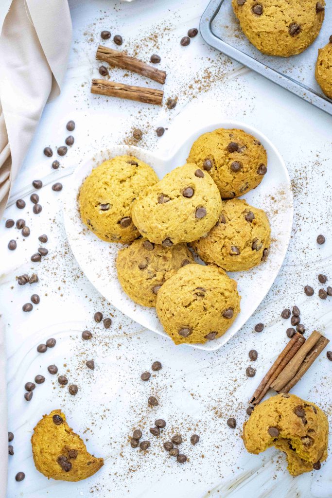A heart-shaped plate holds pumpkin cookies with chocolate chips on a white surface, surrounded by scattered chocolate chips, cinnamon sticks, and cookie crumbs.