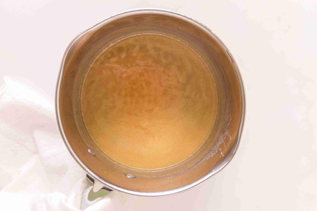 A stainless steel pot filled with a light brown liquid, likely part of a delicious butterscotch sauce recipe, is viewed from above as it sits on a white surface with a white cloth partially visible to the side.
