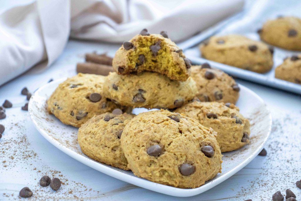 A plate of pumpkin cookies with chocolate chips sits on a white surface, with one cookie broken in half on top. Chocolate chips and cookie crumbs are scattered around, hinting at a deliciously soft pumpkin cookie recipe.