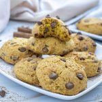 A plate of pumpkin cookies with chocolate chips sits on a white surface, with one cookie broken in half on top. Chocolate chips and cookie crumbs are scattered around, hinting at a deliciously soft pumpkin cookie recipe.