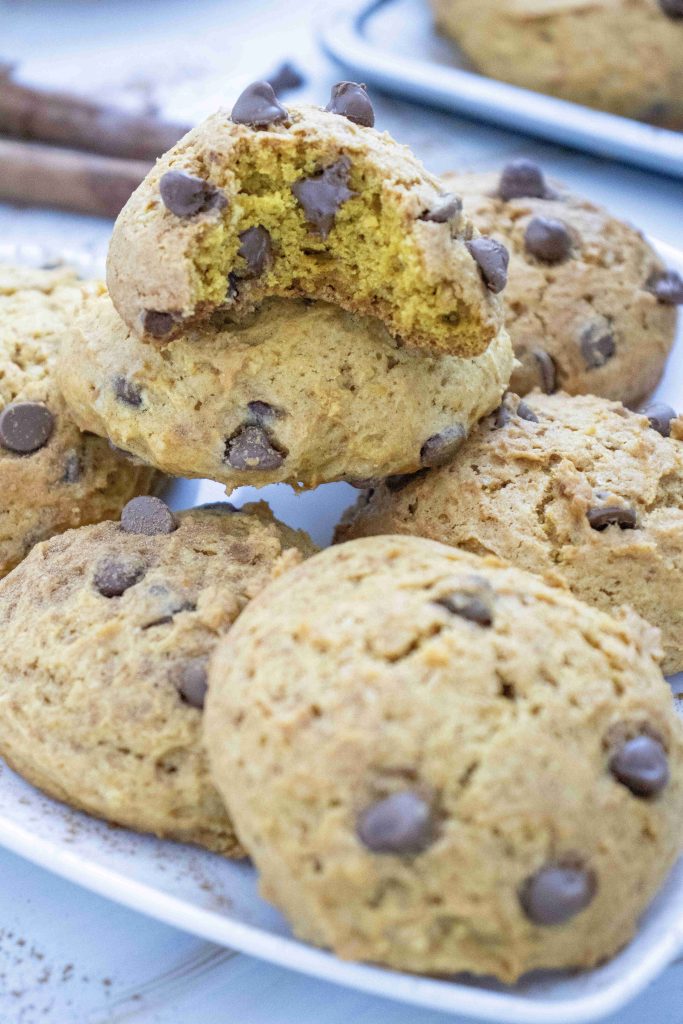 A close-up of several pumpkin cookies with chocolate chips on a plate, with one cookie on top that has a bite taken out of it, showing its soft interior and rich chocolate chips—perfect for anyone looking for a soft pumpkin cookie recipe.