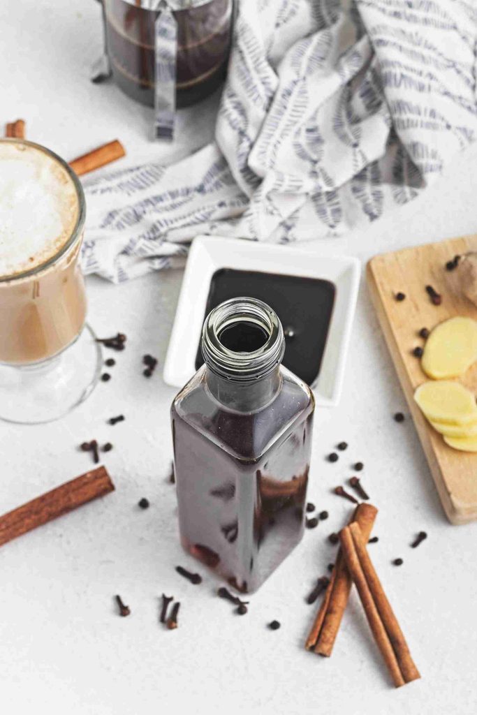 A bottle of dark gingerbread syrup sits on a table with cinnamon sticks, cloves, sliced ginger, a cup of latte, a small dish of syrup, and a striped cloth in the background.