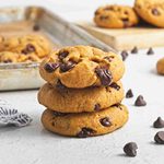 Three vegan pumpkin chocolate chip cookies are stacked in the foreground, with more cookies and chocolate chips scattered around. A baking tray with additional cookies is in the background on a white surface.