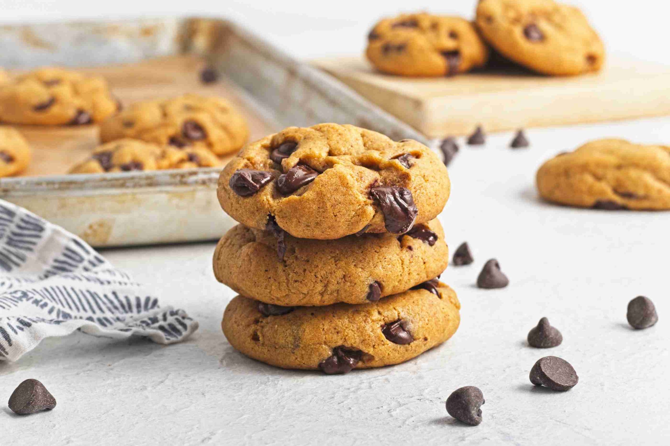 Three vegan pumpkin chocolate chip cookies are stacked in the foreground, with more cookies and chocolate chips scattered around. A baking tray with additional cookies is in the background on a white surface.