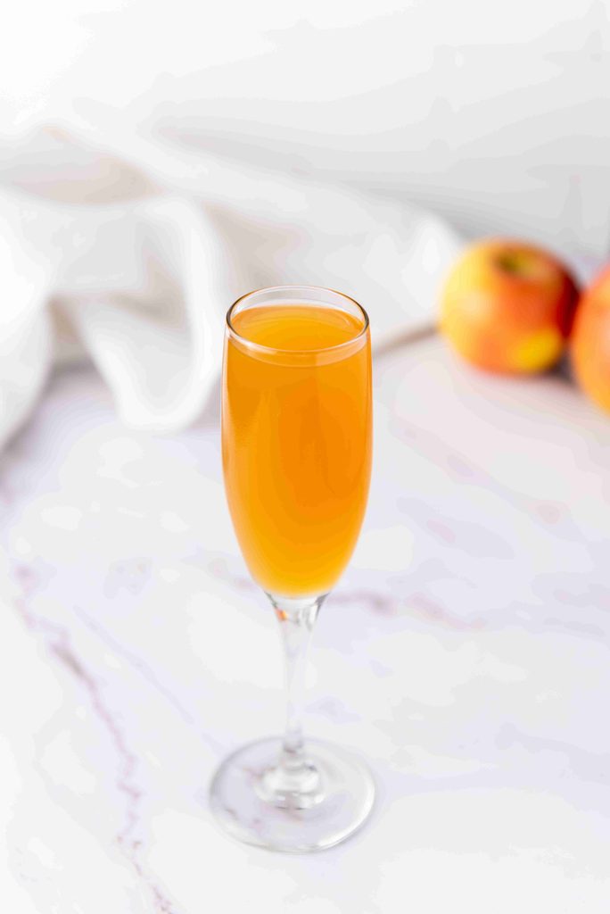 A glass of orange-colored drink, inspired by a fall mimosa recipe, sits elegantly in a champagne flute on a white marble surface, with two apples and a white cloth in the blurred background.