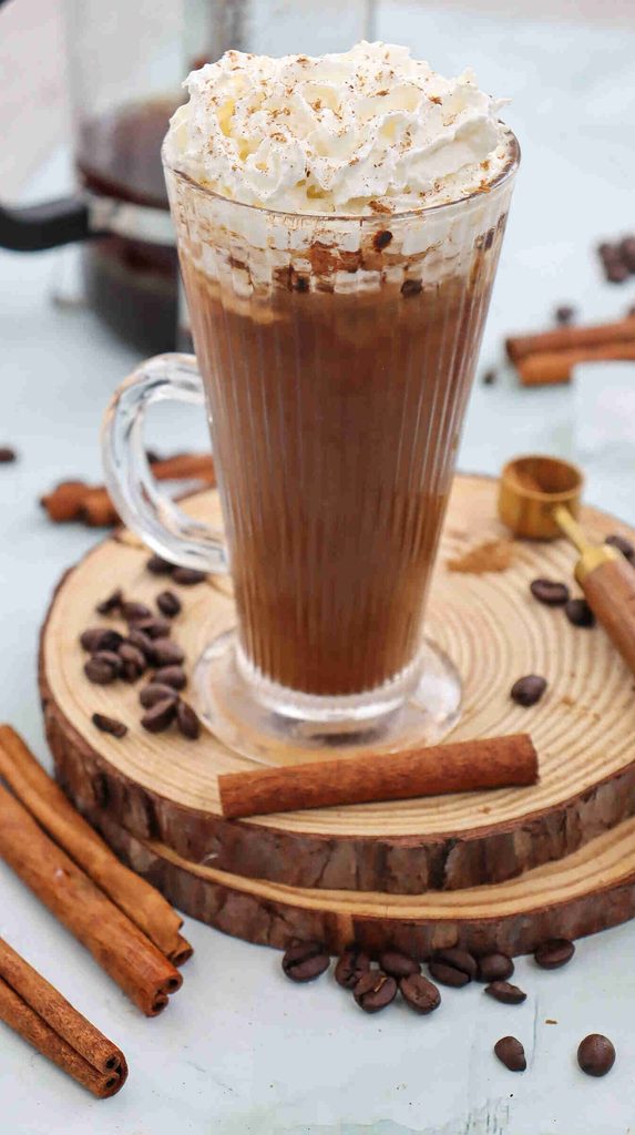 A glass mug of Fall Iced Coffee topped with whipped cream and sprinkled cinnamon sits on a wooden coaster. Coffee beans and cinnamon sticks are scattered around on a light surface.
