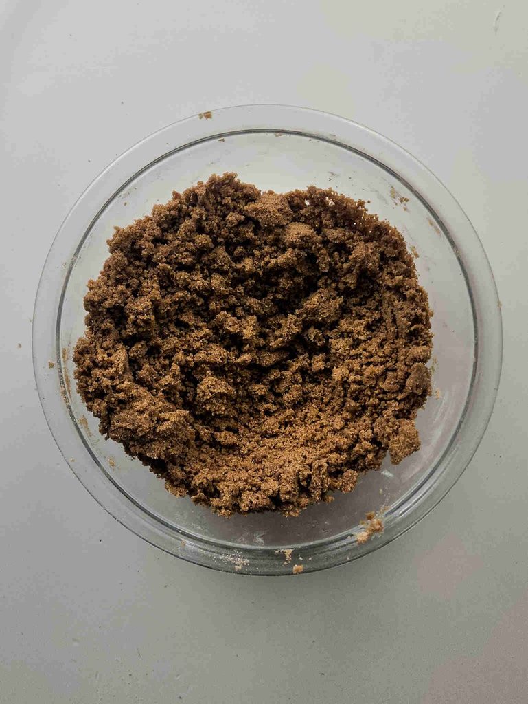 A clear glass bowl filled with a brown, crumbly mixture sits on a light surface. The coarse, grainy texture suggests it could be the topping for pumpkin coffee cake cookies or another autumn-inspired treat.