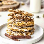 Four iced Pumpkin Coffee Cake Cookies are stacked on a white plate with two cinnamon sticks in front. The top cookie, drizzled with vanilla glaze, is broken in half. A glass of milk and more cookies blur in the background.