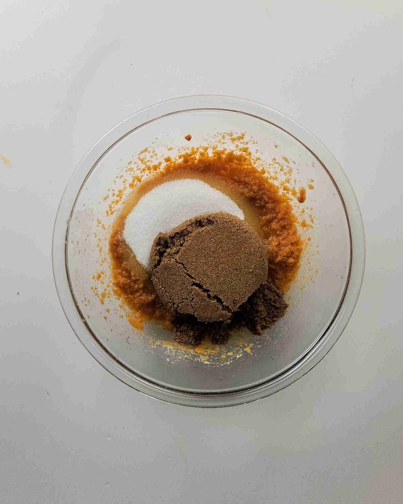 A glass bowl with light brown batter for pumpkin coffee cake cookies, topped with a mound of brown sugar and a pile of white sugar, sits on a white surface.