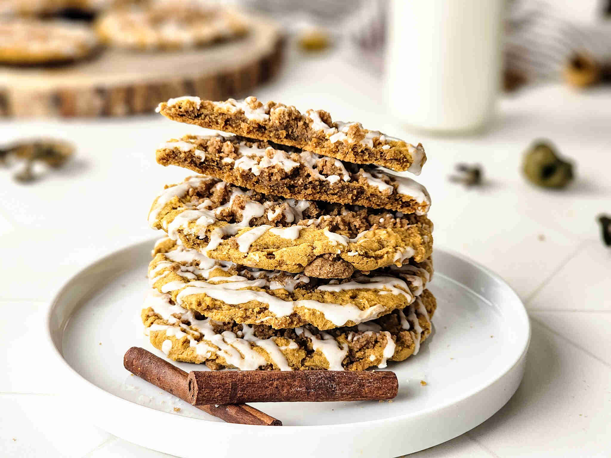 Four iced Pumpkin Coffee Cake Cookies are stacked on a white plate with two cinnamon sticks in front. The top cookie, drizzled with vanilla glaze, is broken in half. A glass of milk and more cookies blur in the background.