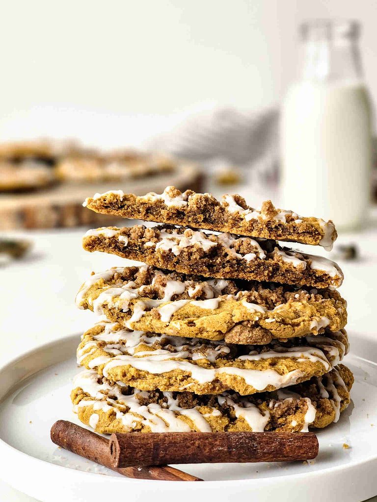 A stack of pumpkin coffee cake cookies with crumb topping sits on a white plate next to cinnamon sticks. A blurred glass of milk is in the background.