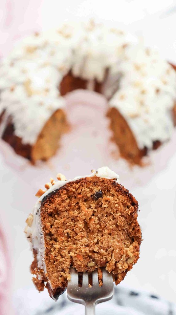 A close-up of a fork holding a slice of spiced fall bundt cake with white icing and chopped nuts, with the whole cake blurred in the background.