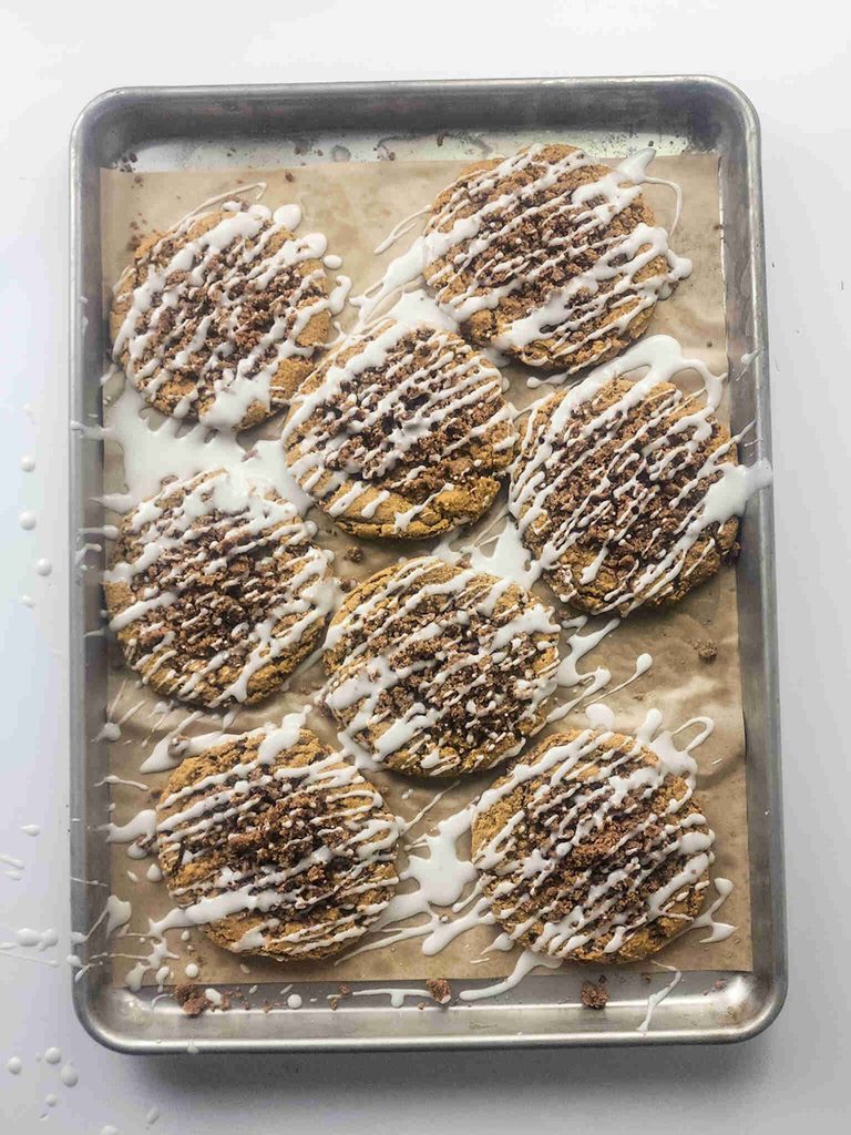 A baking sheet with nine large pumpkin cookies with icing, drizzled and sprinkled with crumbs, rests on parchment paper. Some icing has dripped onto the tray and the surface below.