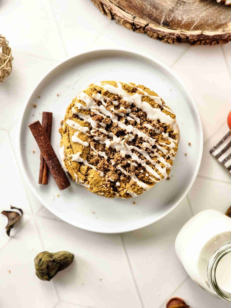A round pumpkin coffee cake with white icing and crumb topping sits on a white plate, reminiscent of pumpkin coffee cake cookies, garnished with cinnamon sticks, beside milk and rustic decor.