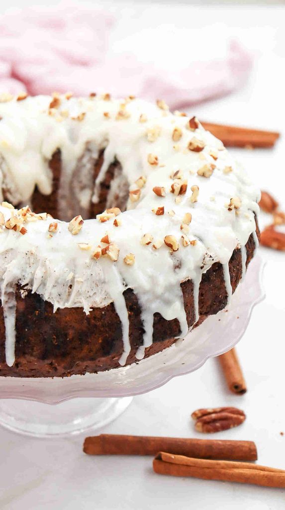 A fall bundt cake with white icing drips, topped with chopped pecans, sits on a glass stand. Cinnamon sticks and pecans are scattered around on a white surface, making this fall bundt cake recipe perfect for cozy autumn gatherings.