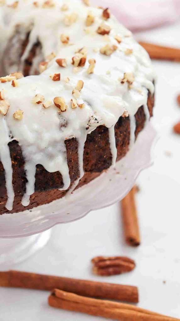 A fall bundt cake with white icing drips and chopped nuts on top sits on a glass stand, surrounded by cinnamon sticks and pecans—a picture-perfect centerpiece for any autumn gathering and the ultimate fall bundt cake recipe.