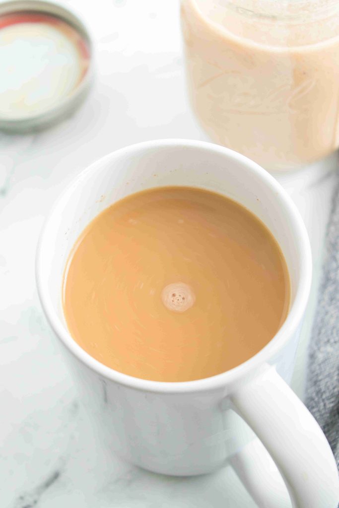 A white mug filled with creamy coffee sits on a white surface, the rich aroma hinting at a touch of fall coffee creamer. A jar with a beige liquid, its lid off—perhaps the homemade creamer itself—is partially visible in the background. A soft gray cloth is nearby.