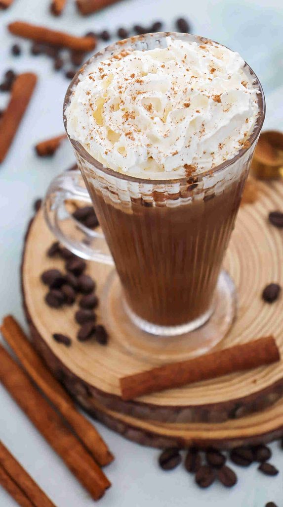 A glass mug of fall iced coffee topped with whipped cream and sprinkled cinnamon sits on a wooden coaster, surrounded by cinnamon sticks and coffee beans.