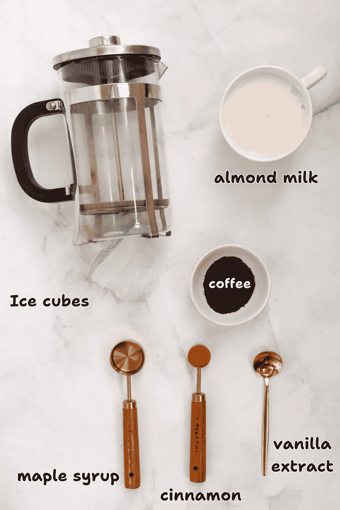 Overhead view of coffee ingredients on a marble surface: a French press, a cup of almond milk, a bowl of coffee grounds, and three measuring spoons labeled for maple syrup, cinnamon, and vanilla extract. Ice cubes are scattered throughout—everything you need to craft the perfect Fall Iced Coffee at home.