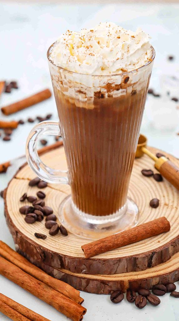 A glass mug of Fall Iced Coffee topped with whipped cream, placed on a wooden coaster, surrounded by coffee beans and cinnamon sticks.